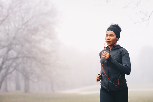 A woman merrily jogging on a misty day in nature. She is smiling. She is wearing headphones, sports clothes and a hairband.