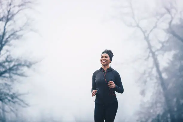 Photo of A woman jogging in the park on a misty day. She is smiling. She wears headphones and sports clothes.