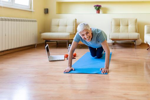 Mature woman exercising at home, looking for new exercise online on laptop.