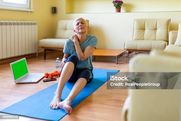 Senior Woman In Sportswear Sitting On Yoga Mat And Massaging Her Shoulder Stock Photo - Download Image Now