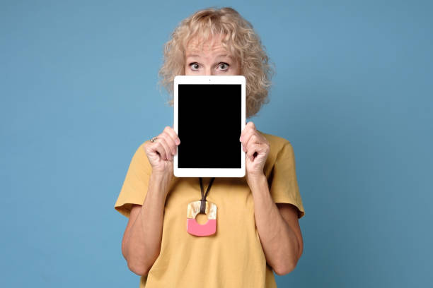 mature woman hiding his face behind portable computer device on blue background - advertisement advertise businesswoman peeking imagens e fotografias de stock