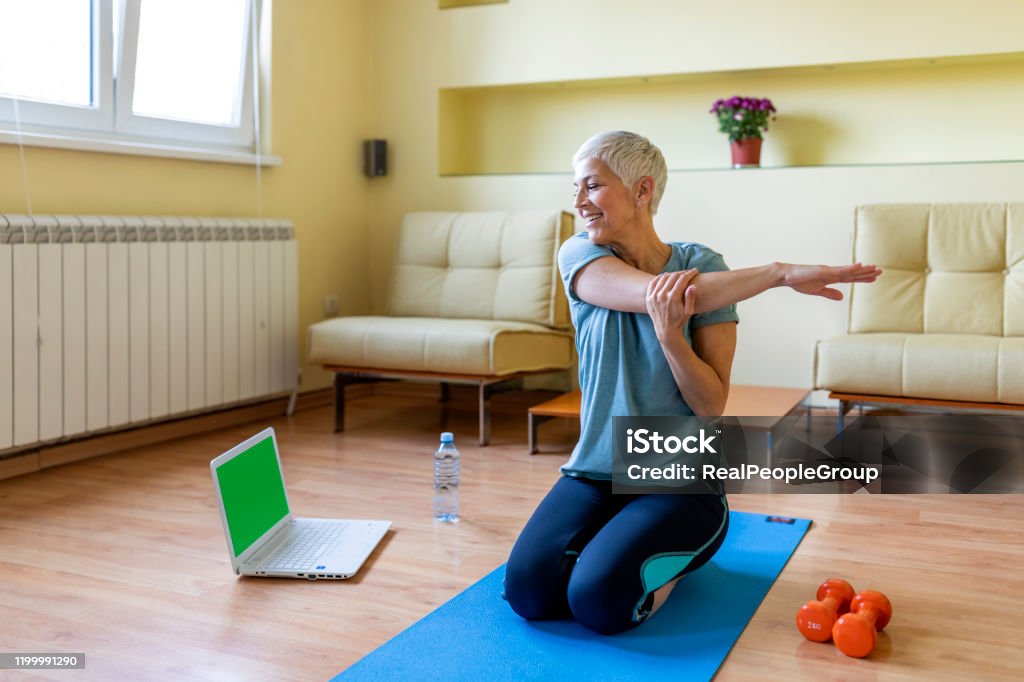 Senior woman exercising in home gym. Beautiful senior woman doing stretching exercise while sitting on yoga mat at home. Mature woman exercising in sportswear by stretching forward to touch toes. Healthy active lady doing yoga. Exercising Stock Photo