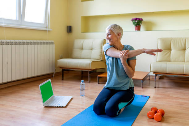 Senior woman exercising in home gym. Beautiful senior woman doing stretching exercise while sitting on yoga mat at home. Mature woman exercising in sportswear by stretching forward to touch toes. Healthy active lady doing yoga. grey hair on floor stock pictures, royalty-free photos & images