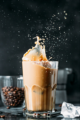 Splash of ice coffee drink on a dark background. Refreshing Iced cappuccino liquid drink pouring into a tall glass with ice cubes. Cold beverage wave. Close-up design liquor milk, coffee and ice.