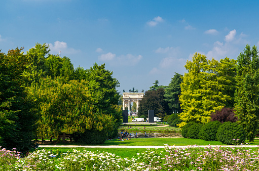 Arch of Peace Arco della Pace gate and green trees, flowers, grass lawn in Parco Sempione park with blue sky background in Milan city centre, Lombardy, Italy