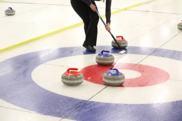 A curler sweeping a red handled curling stone into the button of the house with blue and red handled curling stones.
