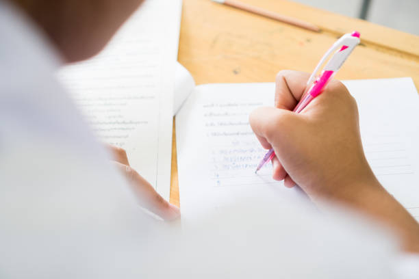 istock Education test concept : Hands student holding pen for testing exams writing answer sheet or exercise for taking fill in exam carbon paper computer on wood table at classroom in high school 1200050724