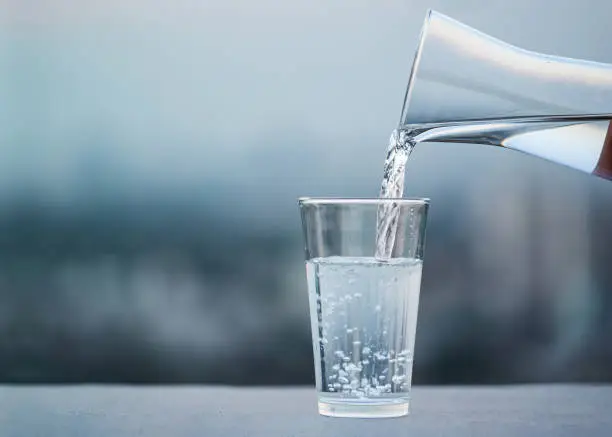Photo of Hand pouring drink water from bottle into glass with blurred city background