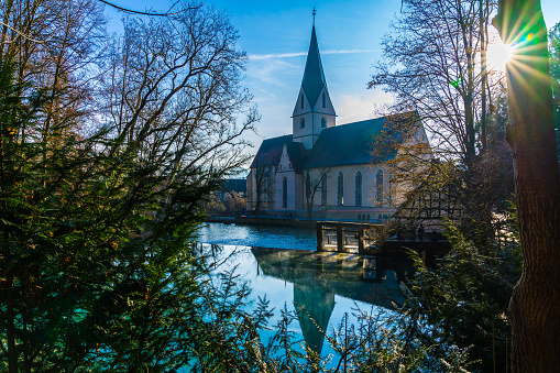 Germany, Early morning fog moving over blue water surface of blue pot or german blautopf in blaubeuren forest reflecting beautiful church building in calm water