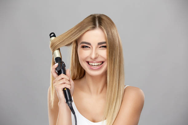beautiful blonde woman using curling iron for her healthy long hair and smiling to camera stock photo