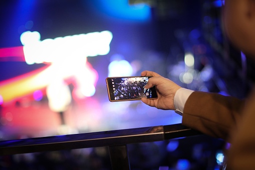 Man use mobile phone shooting video photo of concert in front of stage at night with beautiful blurred bokeh from the lights in background