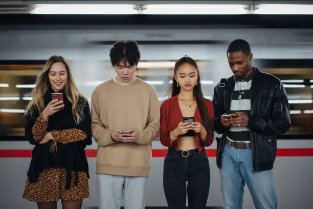 Group of teenage friends, of different ethnicity, is standing in front of train on the move and using cellphones