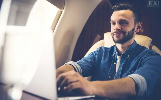 Close-up photo of a happy man in a casual outfit, who is typing something on his laptop and looking at its screen during his flight to another country.