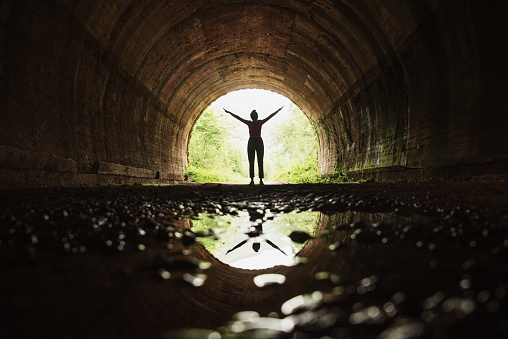 A young woman with arms raised and her reflection on a puddle, inside a dark tunnel with a bright end