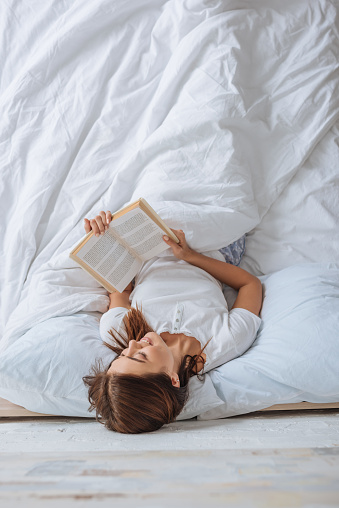 top view of smiling girl reading book while chilling in bed