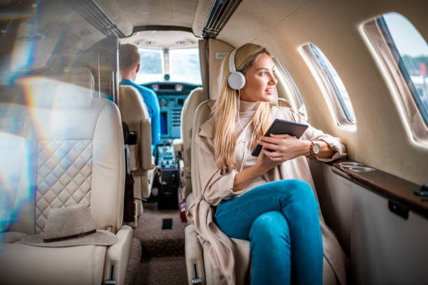 joven mujer de negocios sentada en un jet privado y escuchando música a través de auriculares mientras usa una tableta digital - status symbol audio fotografías e imágenes de stock