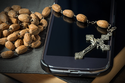Extreme close-up of an old silver crucifix with Jesus Christ and wooden rosary bead on a black smartphone and Holy Bible