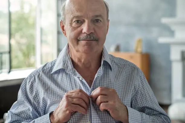 Photo of Senior caucasian hispanic man in the white shirt buttoning shirt up at morning.