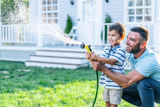 padre spruzza acqua e si diverte con il figlio nel cortile sul retro - annaffiare foto e immagini stock