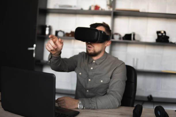 istock Young bearded man sitting against computer with virtual reality glasses trying to touch something or tap some thing in front of what he seeing. Modern technologies. The concept of future technology. 1200095311