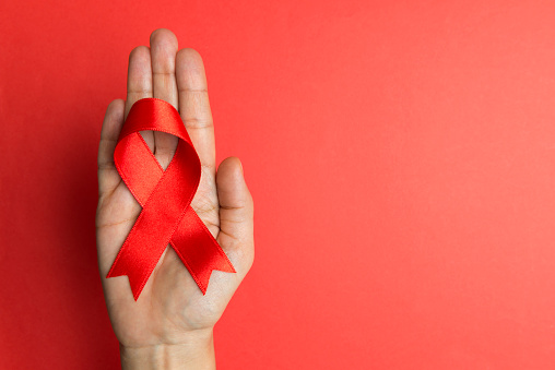 Directly above view of a hand holding red awareness ribbon over red background. 
Some issues for what the red awareness ribbon stands for are AIDS, addiction, alcohol dependence, blood Cancer, heart defects, drug addiction, red ribbon week, Hypertension, Tuberculosis and Zika Virus.