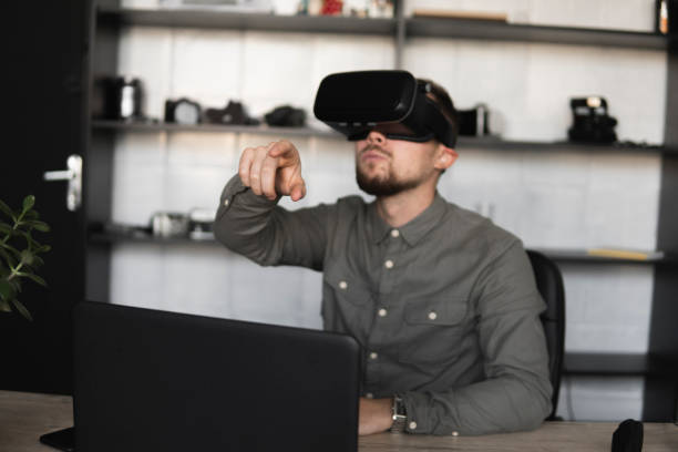 istock Young bearded man sitting against computer with virtual reality glasses trying to touch something or tap some thing in front of what he seeing. Modern technologies. The concept of future technology. 1200105883