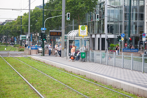 People at tram stop in Dusseldorf in summer captured in street Berliner Allee. A senior woman is standing with other people at stop. Behind is a stop light of street. In background is a modern office building.