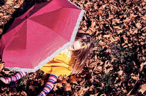 Cute little girl sitting on fall leaves in park.Playful toddler child with red umbrella enjoy in autumn.Playing hide and seek
