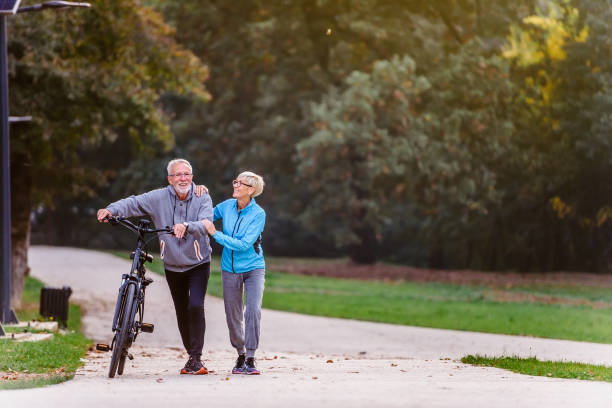 couples aînés actifs gais avec la marche de bicyclette par le stationnement ensemble - cycling senior adult sports helmet men photos et images de collection