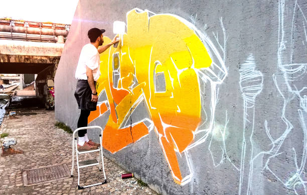 street-künstler arbeiten an farbigen graffiti an öffentlichen raum wand - moderne kunst führen konzept der urbanen kerl malerei live murale mit gelb und orange aerosol farbe spray - helle sonnenfackel filter - straßenmaler stock-fotos und bilder