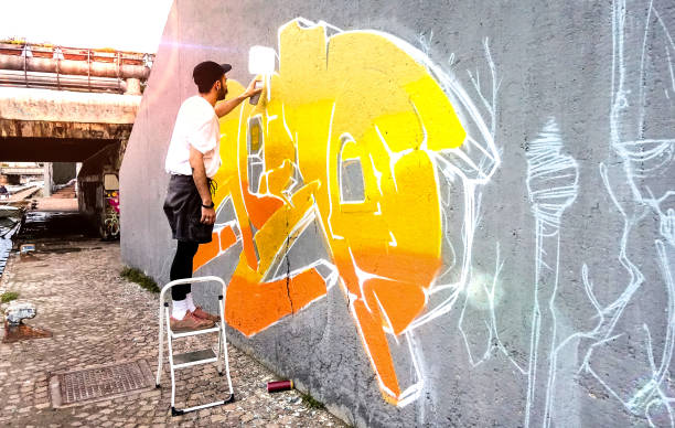 公共スペースの壁で色付きの落書きに取り組むストリートアーティスト - 現代美術は、黄色とオレンジ色のエアロゾルカラースプレーでライブ壁画を描く都市の男のコンセプトを実行します  - paintings art painted image space ストックフォトと画像