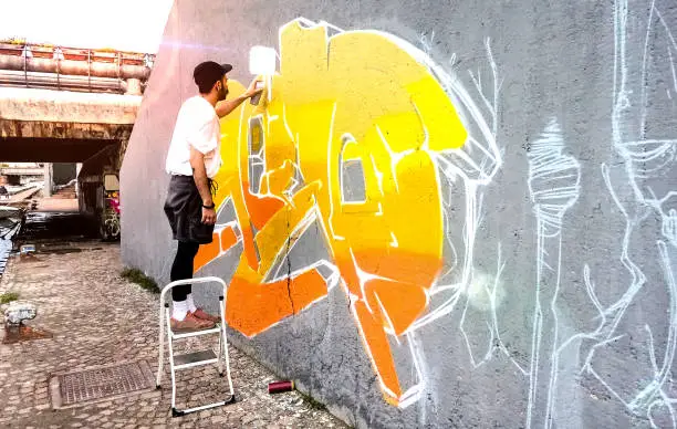 Photo of Street artist working on colored graffiti at public space wall - Modern art perform concept of urban guy painting live murales with yellow and orange aerosol color spray - Bright sunflare filter