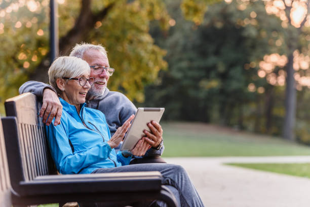 smiling senior active couple sitting on the bench looking at tablet computer - digital tablet women enjoyment happiness imagens e fotografias de stock