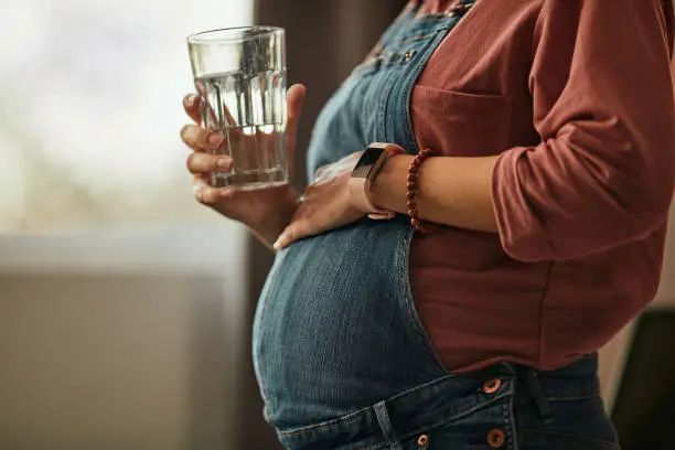 Close up of pregnant caucasian woman standing next to window, touching belly and holding glass of water.