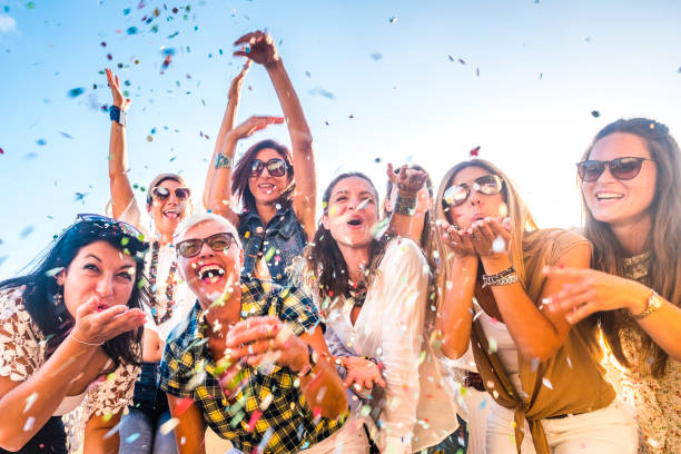 Cheerful group of happy people mixed ages generations women having fun all together during celebration party or carnival - view of fiends blowing. coloured confetti and laugh a lot in friendship Cheerful group of happy people mixed ages generations women having fun all together during celebration party or carnival - view of fiends blowing. coloured confetti and laugh a lot in friendship public celebratory event stock pictures, royalty-free photos & images