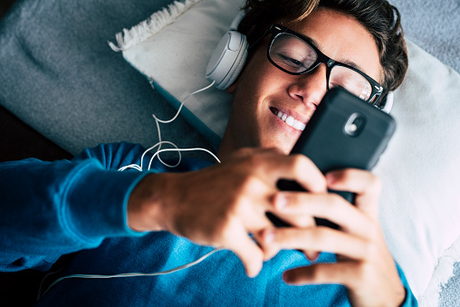 Close up of young phone addicted teenager using mobile cellular lay down on the sofa at home - happy technology millennial people with internet connection device  listening music with heaphones