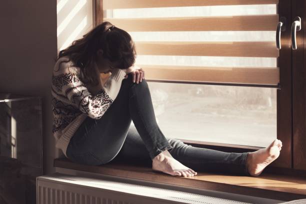 istock Beautiful sad young woman sitting on the window 1200159054