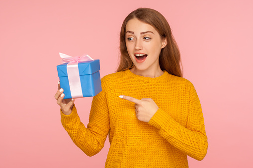Wow, look at my gift. Portrait of astonished red hair girl in sweater pointing at present box and looking with amazement, surprised by holiday present. indoor studio shot isolated on pink background
