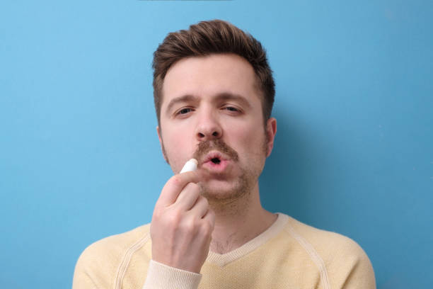 istock Guy applying balm on cracked dry lips using hygienic balm isolated on blue background 1200171153