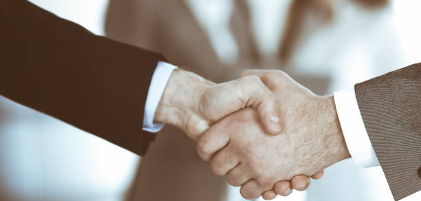 istock Business people shaking hands while standing with colleagues after meeting or negotiation, close-up. Group of unknown businessmen and women in modern office. Teamwork, partnership and handshake concept, toned picture 1200175337