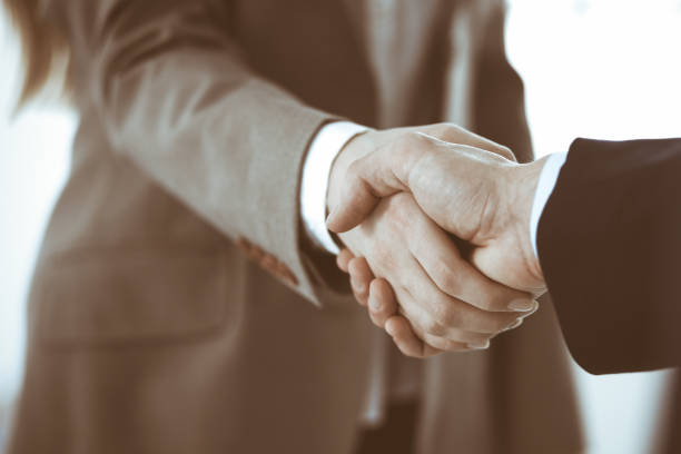 istock Business people shaking hands while standing with colleagues after meeting or negotiation, close-up. Group of unknown businessmen and women in modern office. Teamwork, partnership and handshake concept, toned picture 1200175341