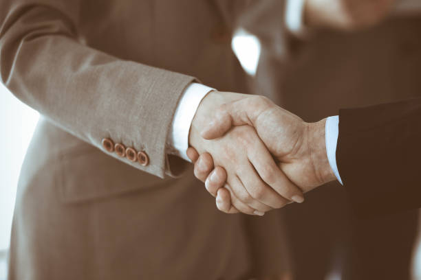 istock Business people shaking hands while standing with colleagues after meeting or negotiation, close-up. Group of unknown businessmen and women in modern office. Teamwork, partnership and handshake concept, toned picture 1200175349