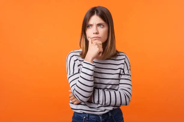 Portrait of worried frustrated woman with brown hair in long sleeve striped shirt standing, holding her chin and frowning, thinking with serious look. indoor studio shot isolated on orange background