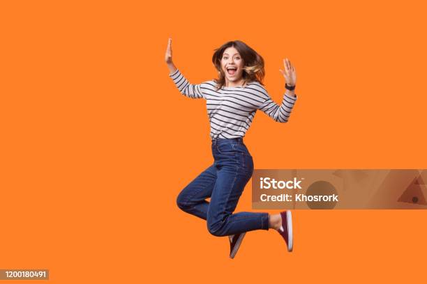 Portrait Of Extremely Excited Pretty Woman With Brown Hair In Long Sleeve Striped Shirt And Denim Jumping Indoor Studio Shot Isolated On Orange Background Stock Photo - Download Image Now
