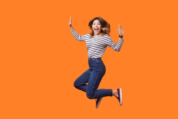 Photo of Portrait of extremely excited pretty woman with brown hair in long sleeve striped shirt and denim jumping. indoor studio shot isolated on orange background