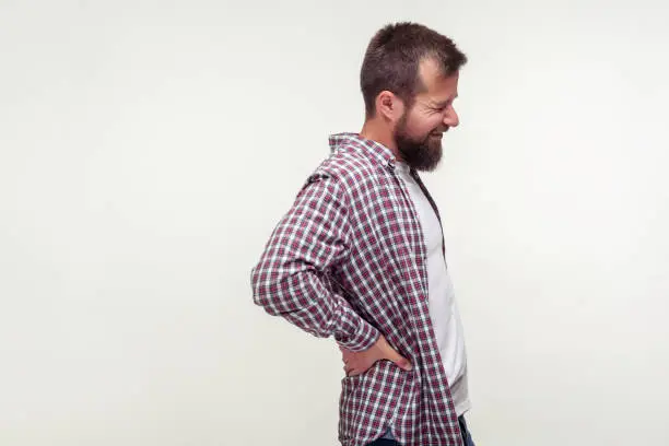 Portrait of unhealthy bearded man in casual plaid shirt touching sore back feeling backpain, pinched nerve aching lower spine, kidney inflammation. indoor studio shot isolated on white background