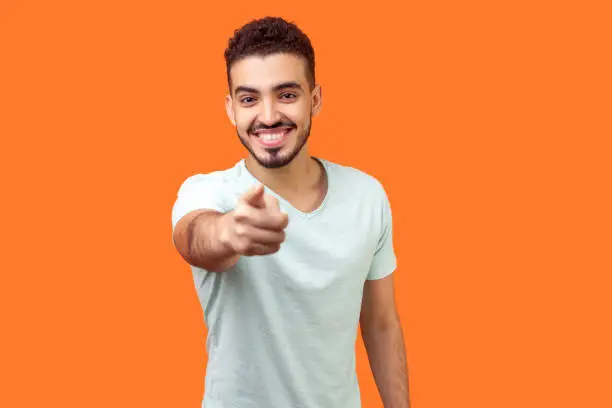 Hey you! Portrait of cheerful handsome brunette man with beard in white t-shirt pointing finger at camera with joyful toothy smile, choosing you. indoor studio shot isolated on orange background