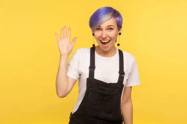 Photo of Portrait of attractive pleased friendly hipster woman waving hand saying hello. isolated on yellow background, studio shot