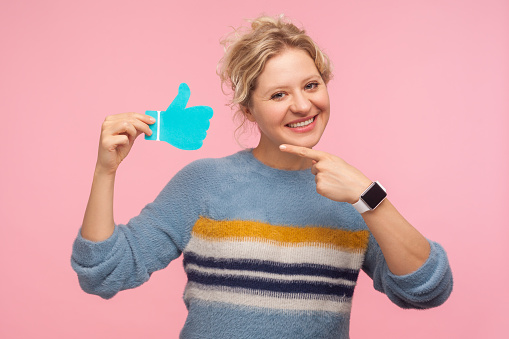Like button. Portrait of cheerful woman in sweater pointing at thumbs up blue icon, follower notification, recommendation and good feedback in social media. studio shot isolated on pink background