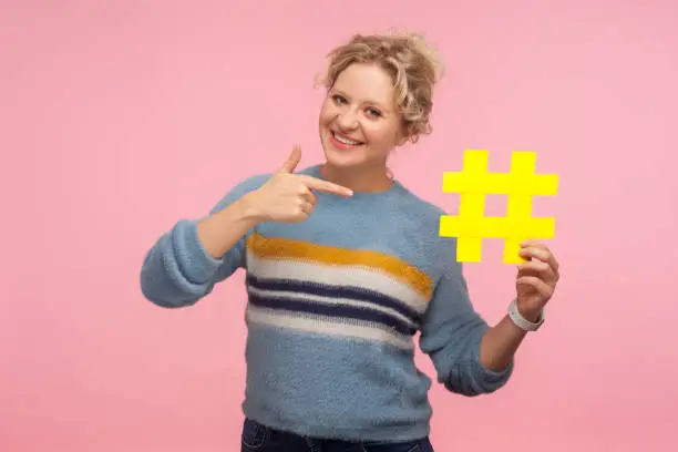 Photo of Look at hashtag. Portrait of cute happy woman with short curly hair in warm sweater pointing at big yellow hash sign